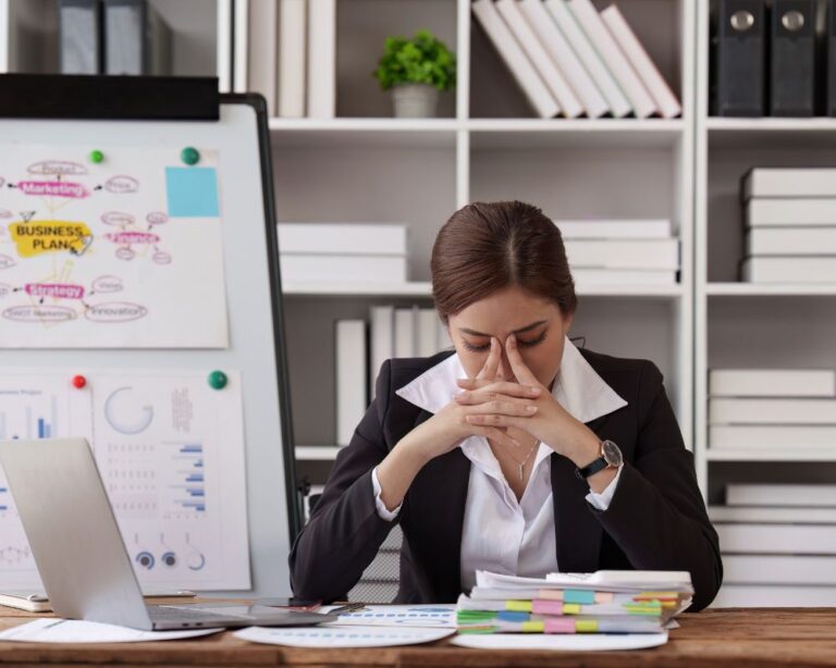 Gestresste Frau sitzt am Schreibtisch mit Whiteboard und Notizen